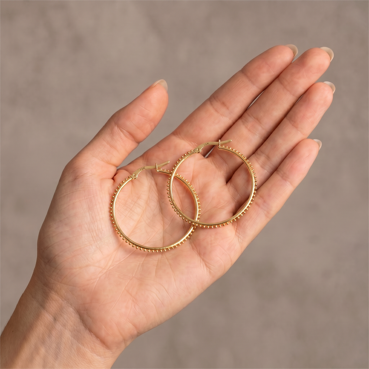 A hand holding a pair of gold hoop earrings against a neutral background.