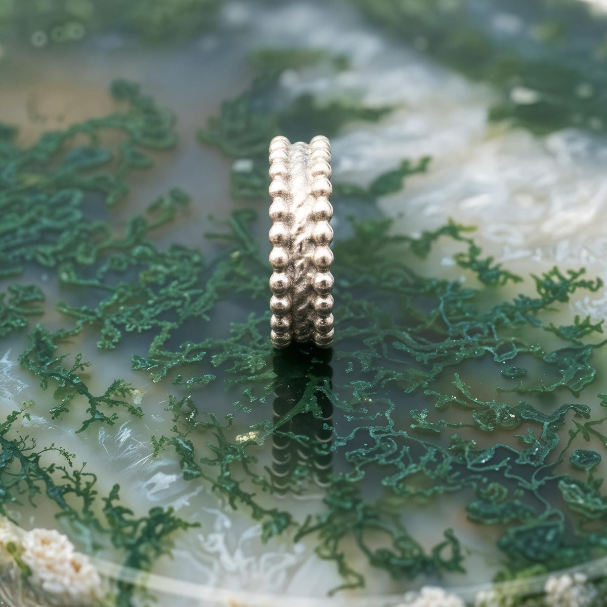 A silver ring with a unique, textured design sits on a surface covered in green moss or algae.