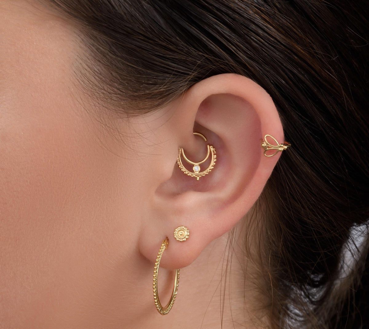 A close-up of a person's ear adorned with multiple gold jewelry pieces, including hoop earrings, a piercing, and a ring.