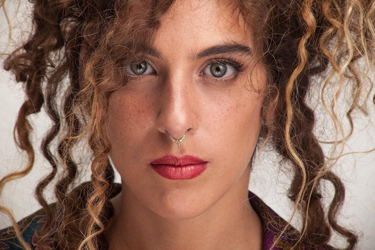 A close-up portrait of a woman with curly hair and a nose piercing, looking directly at the camera with a serious expression.