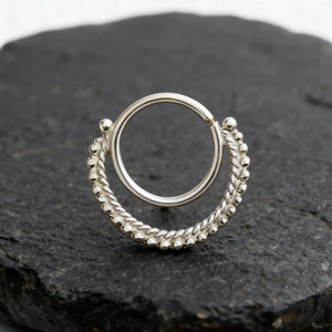 A silver-colored metal ring with a twisted design, resting on a dark surface.