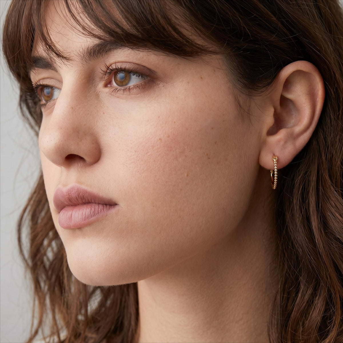 A close-up portrait of a woman with brown hair and a thoughtful expression, wearing gold earrings.