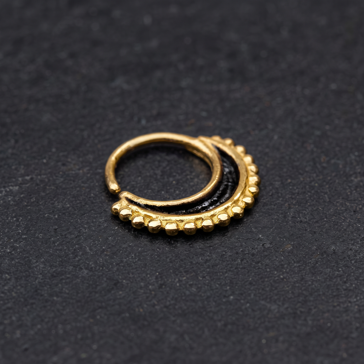 A gold-colored metal nose ring with a black center and decorative elements is shown against a dark background.