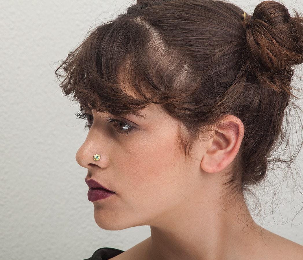 A close-up portrait of a woman with dark hair styled in a bun, wearing a nose ring.