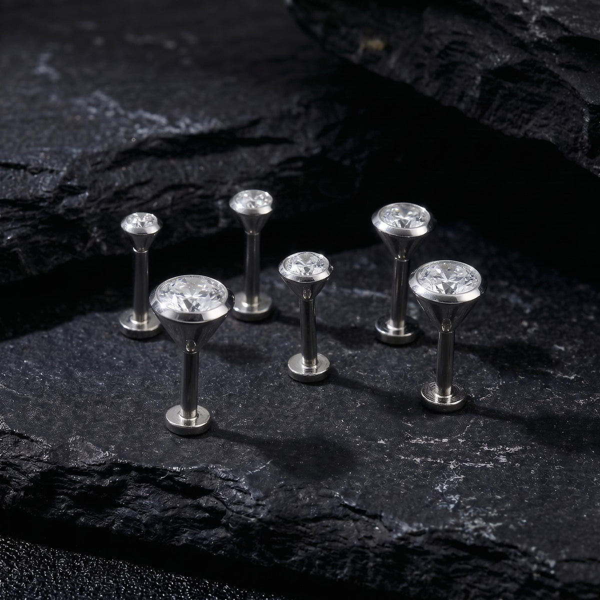 A group of six silver-colored diamond-encrusted earrings or studs are arranged on a dark, textured surface, such as a rock or a black background.