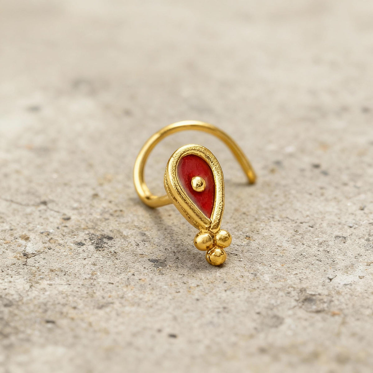 A gold-colored nose ring with a red gemstone is shown against a light-colored background.