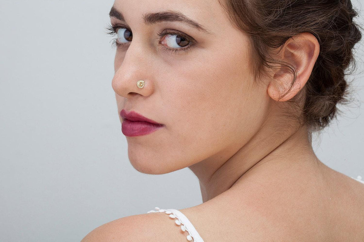 A close-up portrait of a woman with brown hair, wearing a white top and a nose ring, looking off to the side with a serious expression.