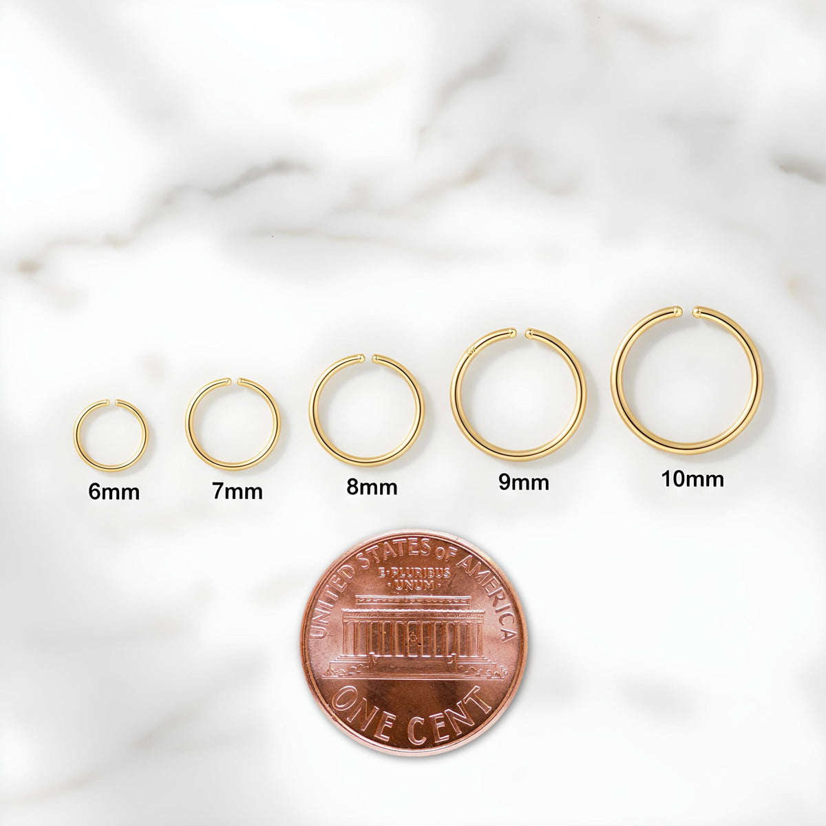 a series of gold-colored rings of different sizes, ranging from 6mm to 10mm, arranged in a row. A penny is placed in front of the rings to provide a scale reference for the sizes.