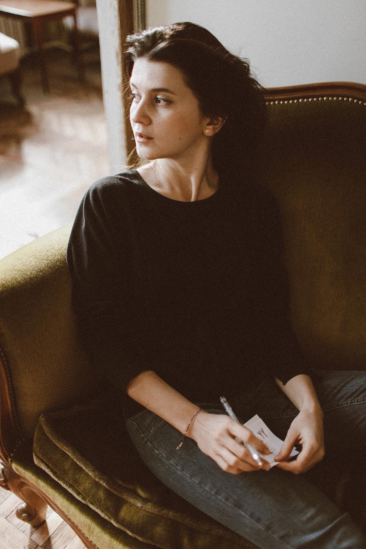 A woman sits on a green couch, holding a pen and paper, with a thoughtful expression on her face.