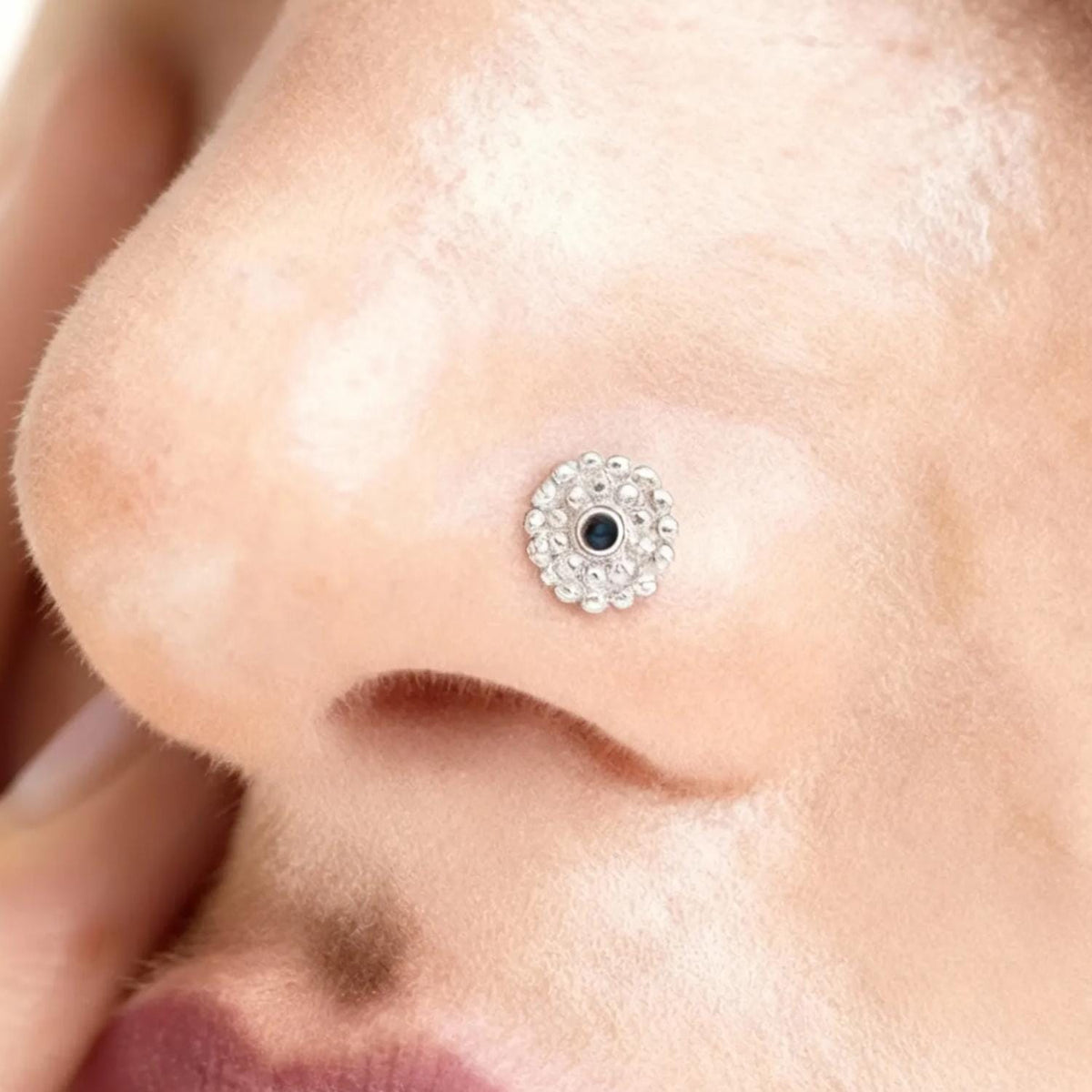 A close-up of a person's nose with a small, ornate nose ring featuring a black center enamel and white metal circle around it.