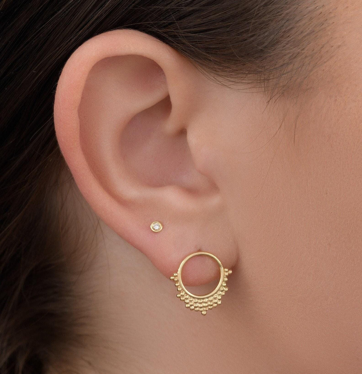 Solid 14k Gold Hoop Stud Earrings: A close-up view of a person's ear adorned with two gold earrings. The earrings have a circular design with a small gemstone in the center, and the person's hair is visible on the side of the ear.