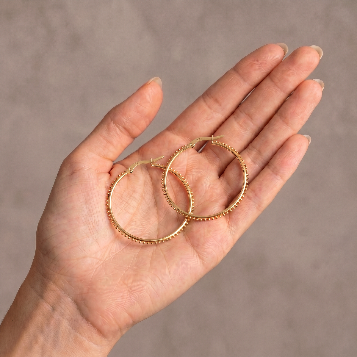 A hand holding a pair of gold hoop earrings against a neutral background.