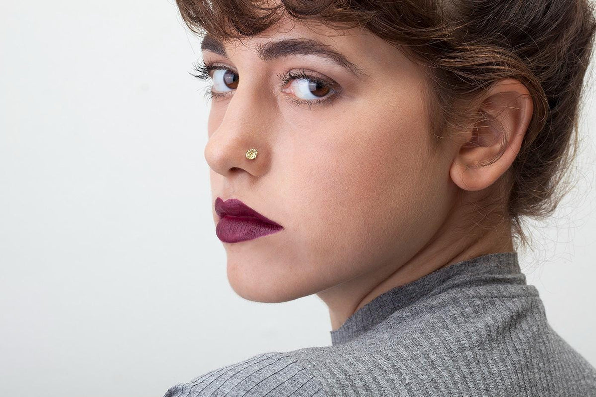 A close-up portrait of a woman with brown hair and a nose piercing, wearing a gray sweater and looking off to the side with a serious expression.