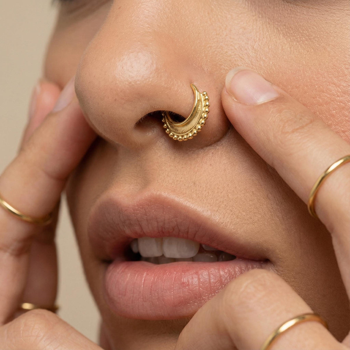 A close-up of a person's face, focusing on their nose and lips, with a gold nose ring and rings on their fingers.