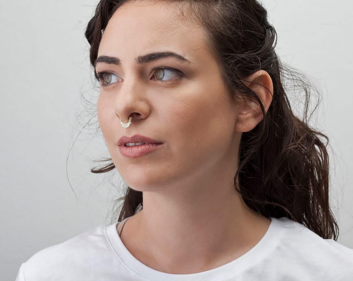 A woman with dark hair and a nose piercing is looking off to the side with a thoughtful expression.