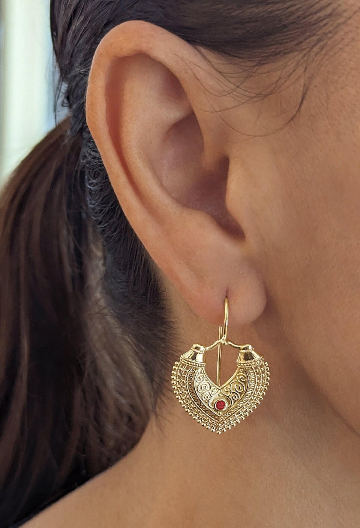 Gold Dangle Earrings: A close-up view of a person's ear wearing a gold earring with a heart-shaped design and a red gemstone.