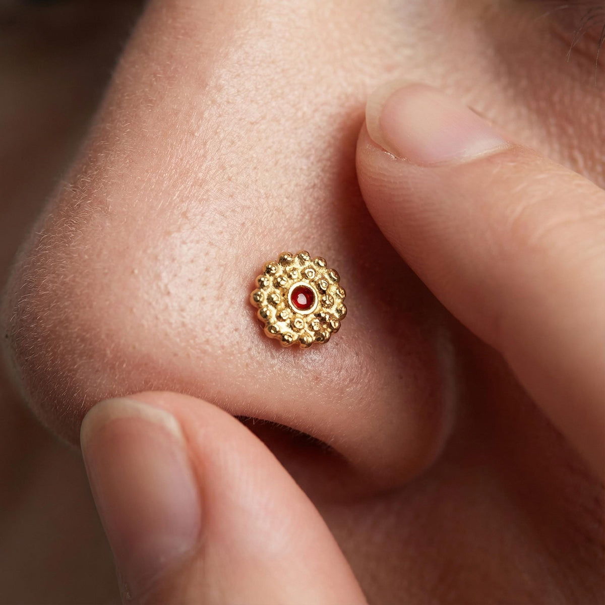A close-up of a person's nose with a small, ornate gold nose ring with a red gemstone in the center.