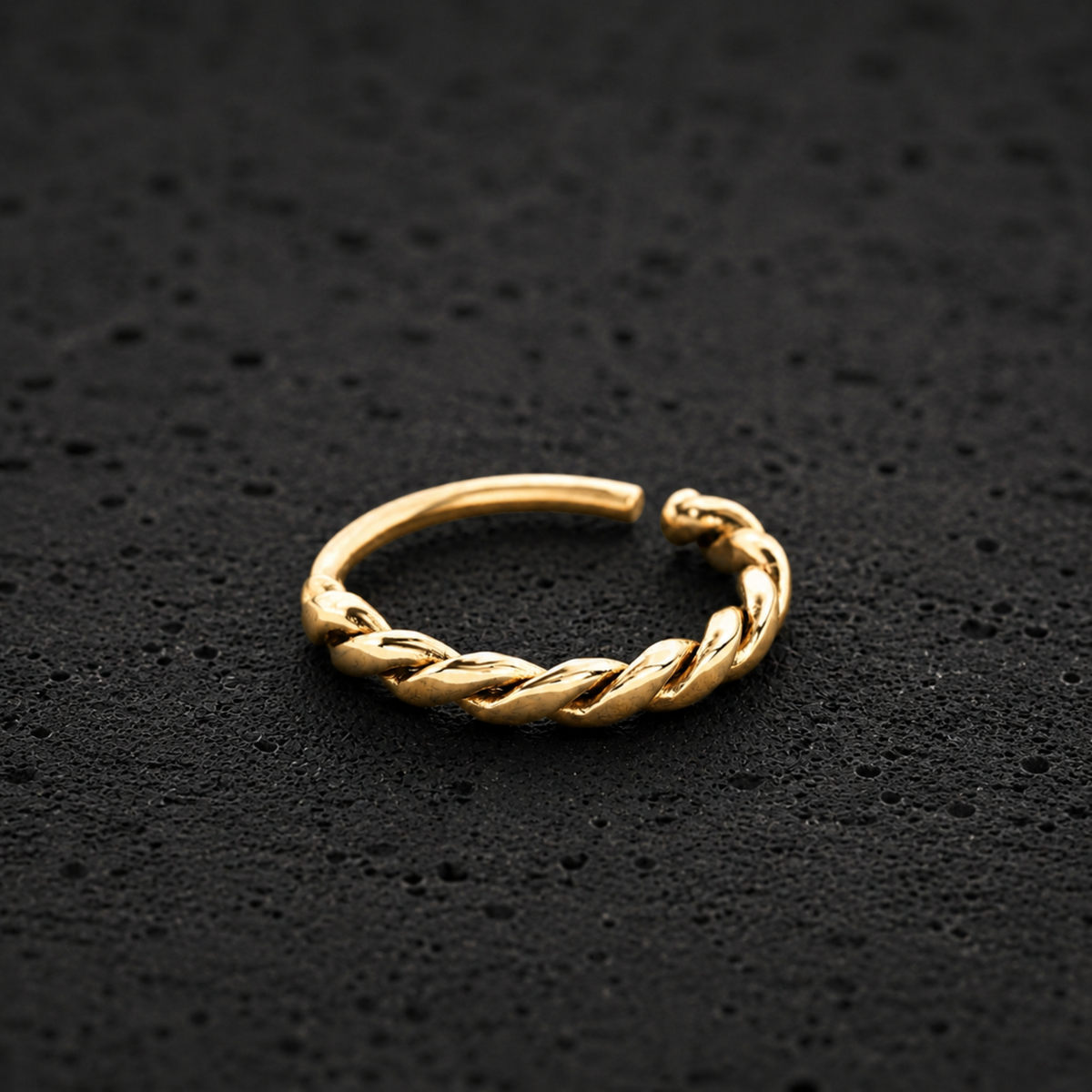 A gold-colored metal ring with a twisted, braided design is shown against a dark, textured background.