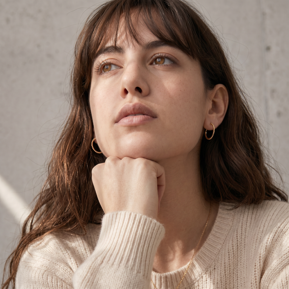 A woman with long brown hair wearing a white sweater and gold hoop earrings looks pensively to the side, her hand resting on her chin.