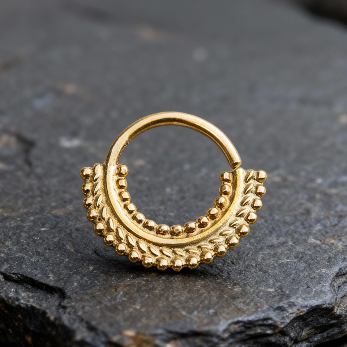 A gold-colored metal nose ring with a decorative leaf-like design is resting on a dark surface.