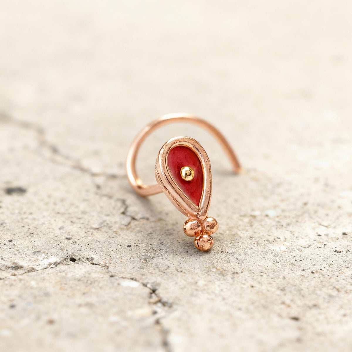 A close-up image of a small, ornate nose ring with a red gemstone and a gold-colored metal frame.