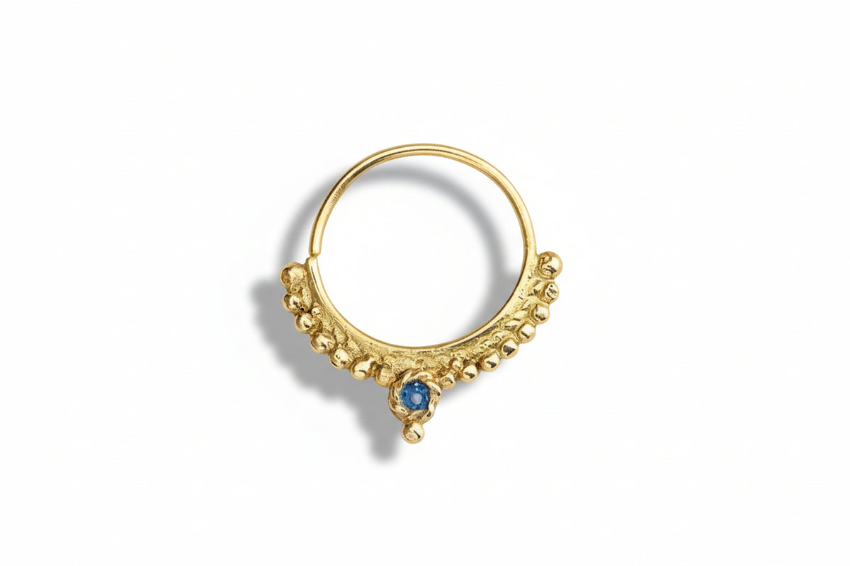 A gold-colored nose ring with a blue gemstone, possibly a sapphire, set in a decorative pattern.
