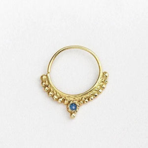 A gold-colored nose ring with a blue gemstone, possibly a sapphire, set in a decorative pattern.