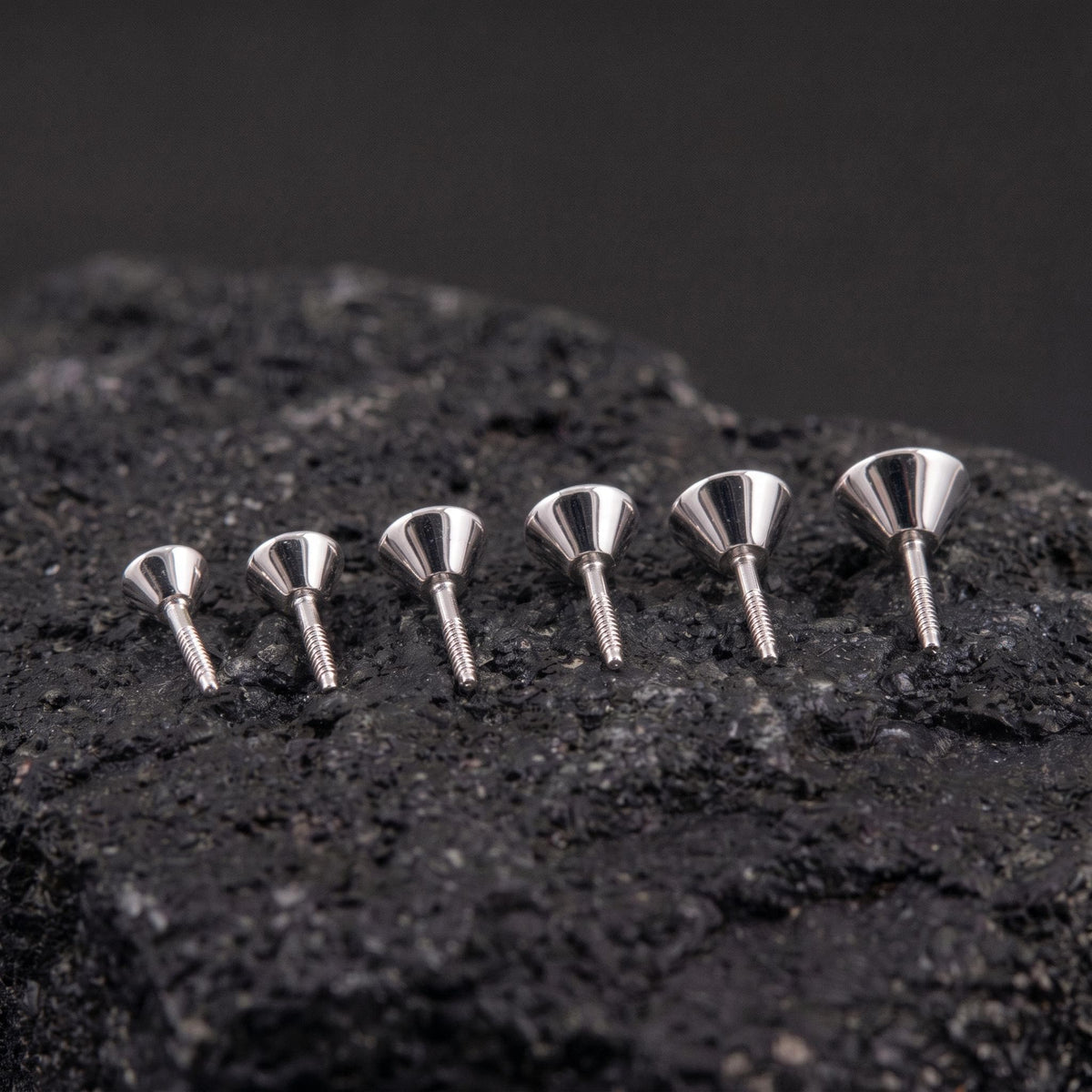 Six silver-colored metal objects are arranged in a row on a dark surface, with their shapes and sizes varying.