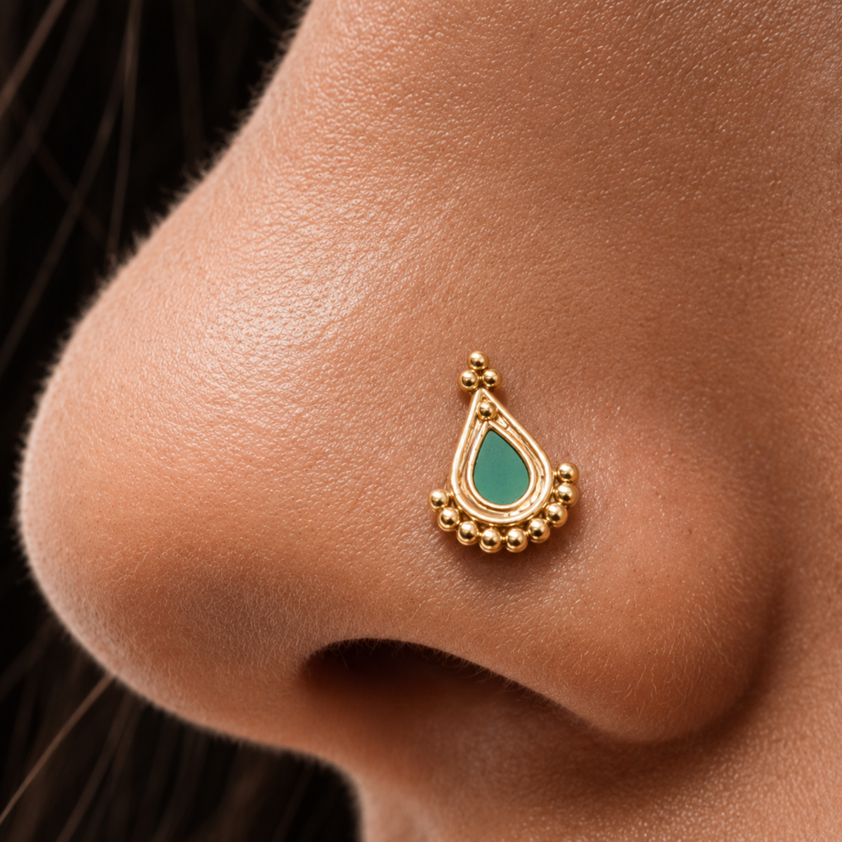 A close-up of a person's nose with a gold-colored nose ring featuring a green stone accent.