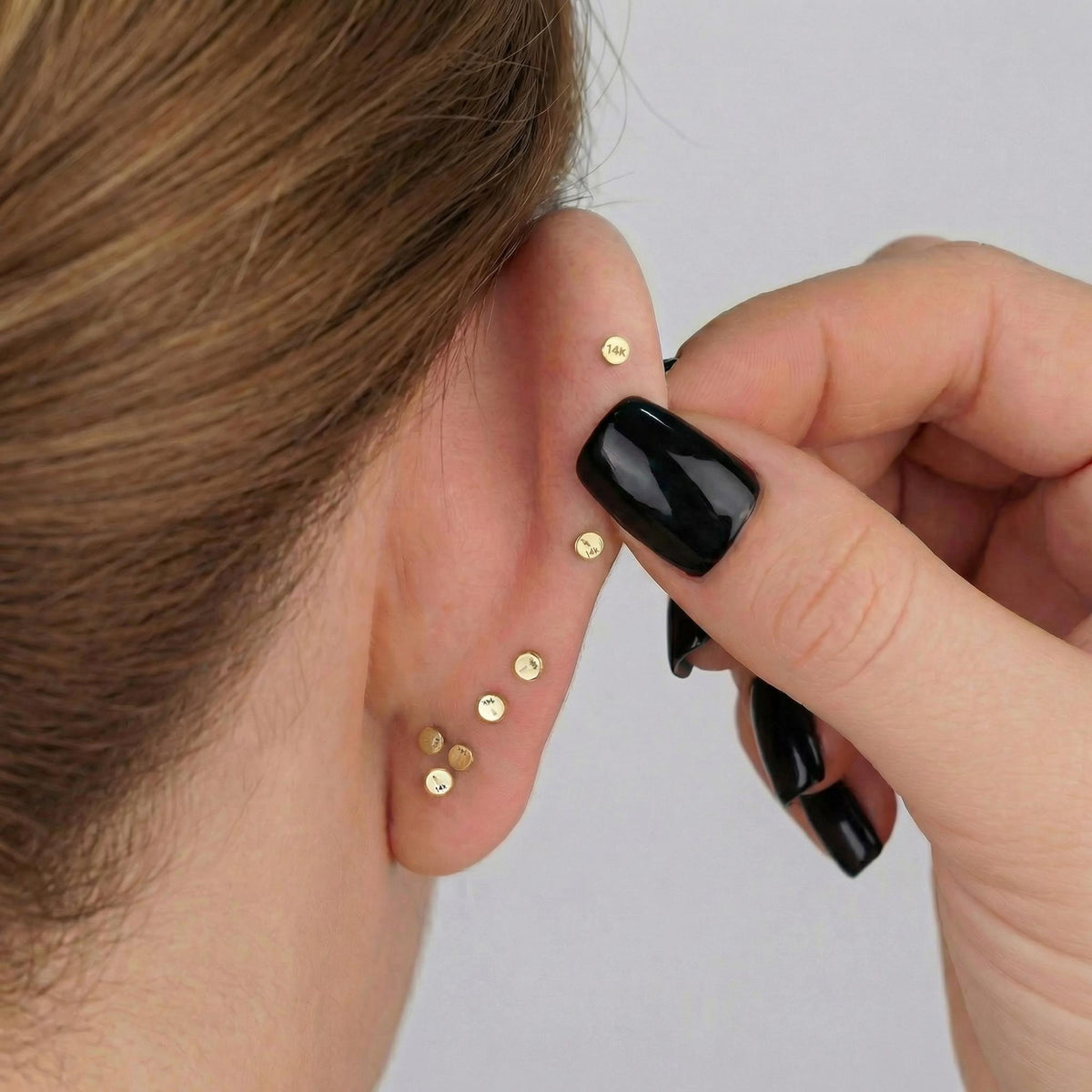 A person's hand is holding a pair of gold earrings with small round stones, with the person's ear being pierced.