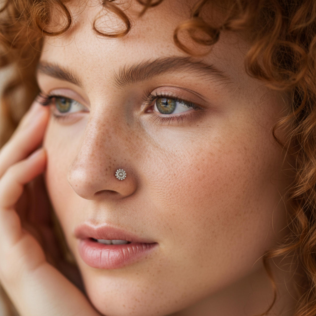 A close-up portrait of a woman with curly red hair, wearing a nose ring, and looking pensively at the camera.