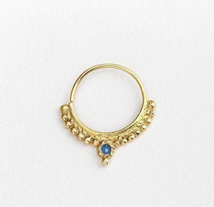 A gold-colored nose ring with a blue gemstone, possibly a sapphire, set in a decorative pattern.