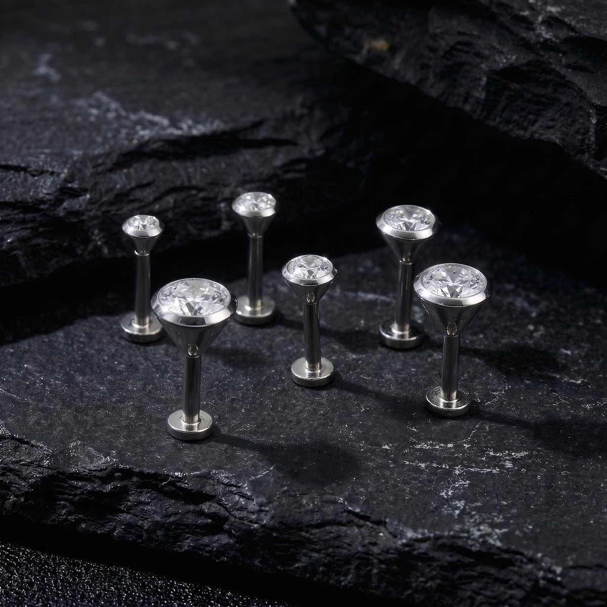 A group of six silver-colored diamond-encrusted earrings or studs are arranged on a dark, textured surface, such as a rock or a black background.