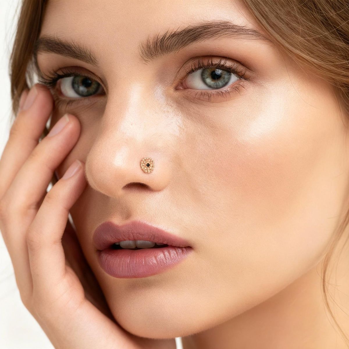 A close-up portrait of a woman with her hand on her face, wearing a nose stud.