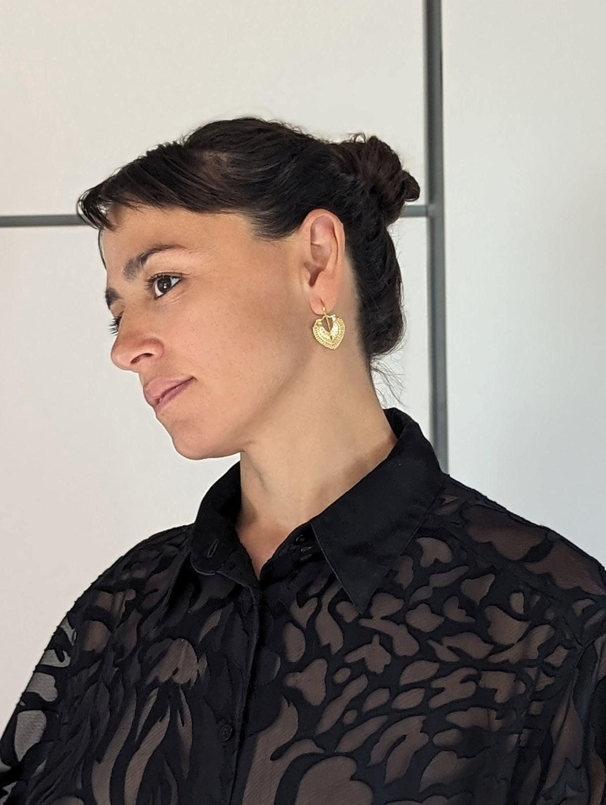 A woman wearing a black patterned blouse and gold earrings is shown in profile, with a neutral expression.