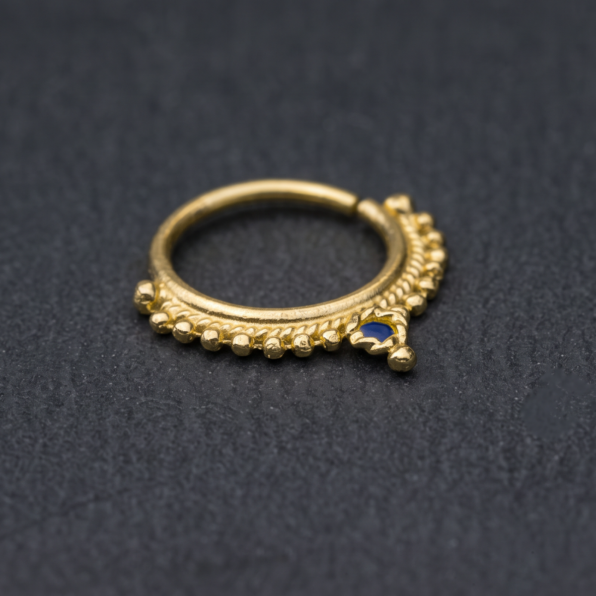A gold-colored nose ring with a blue gemstone is shown against a dark background.