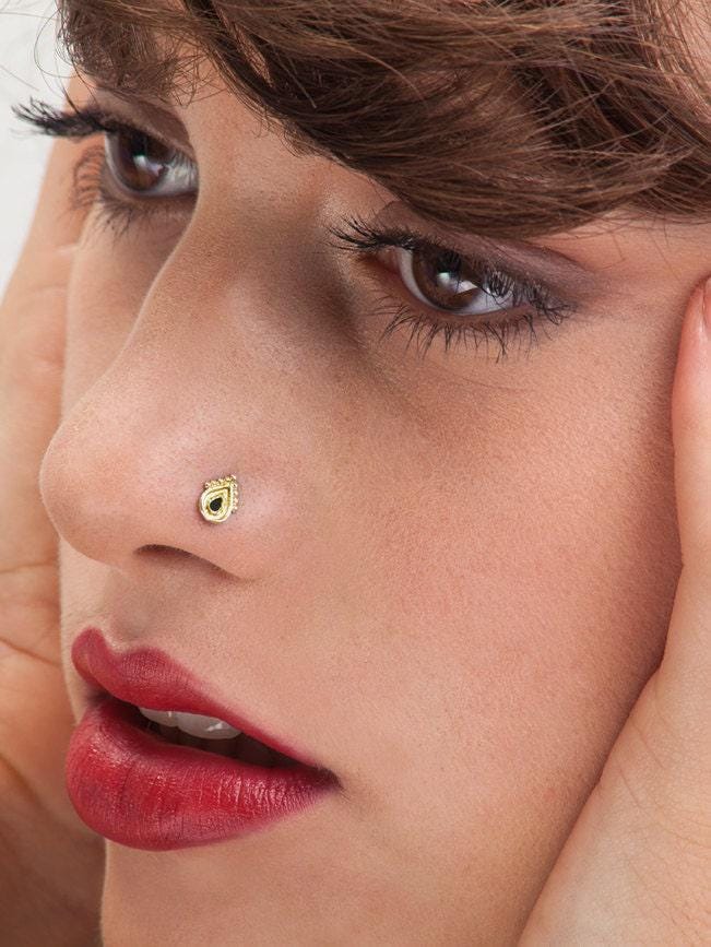 A close-up of a woman's face, focusing on her nose and lips, with her eyes closed. She has dark hair and is wearing a gold nose stud.