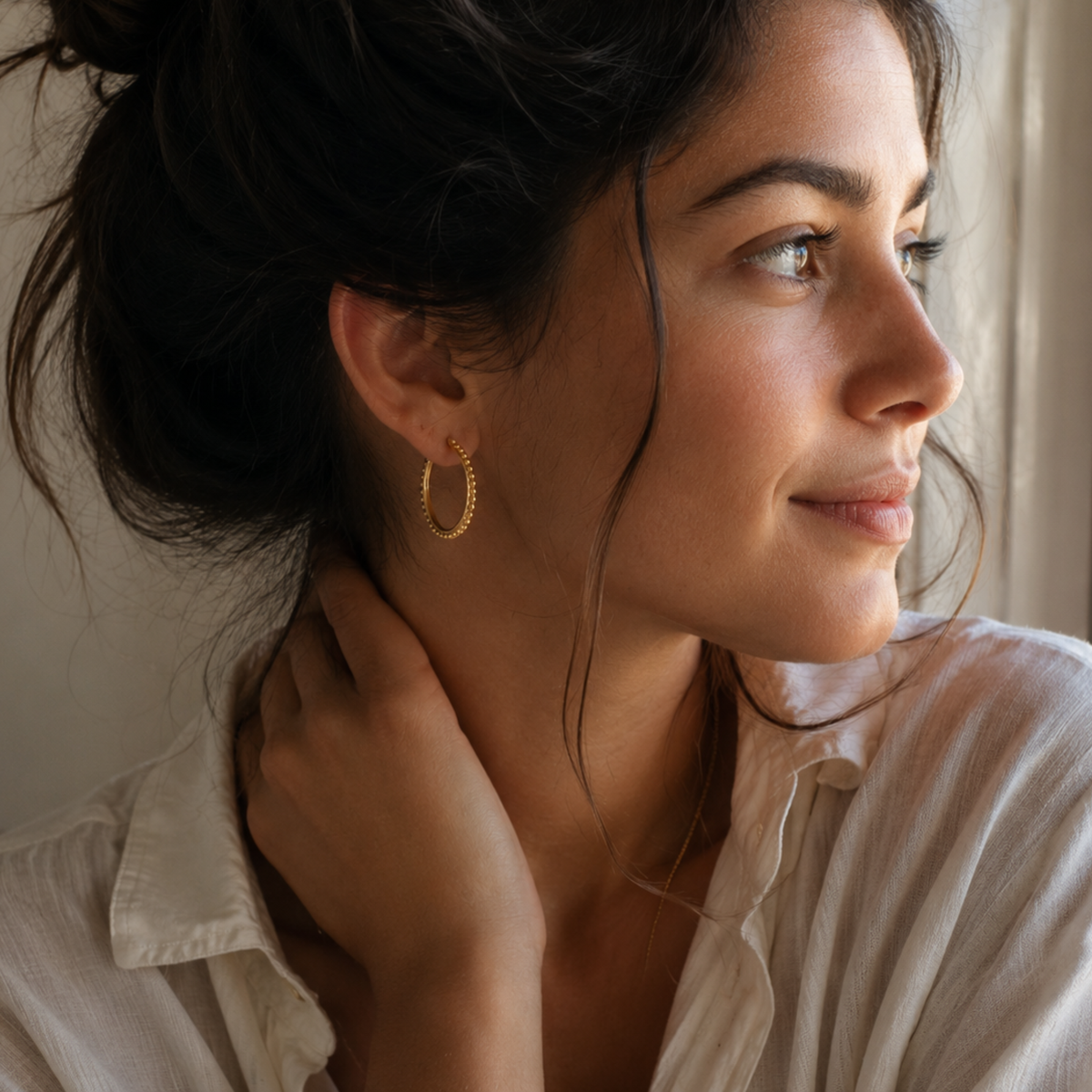 A woman with dark hair and a thoughtful expression gazes out a window, wearing a white shirt and gold hoop earrings.