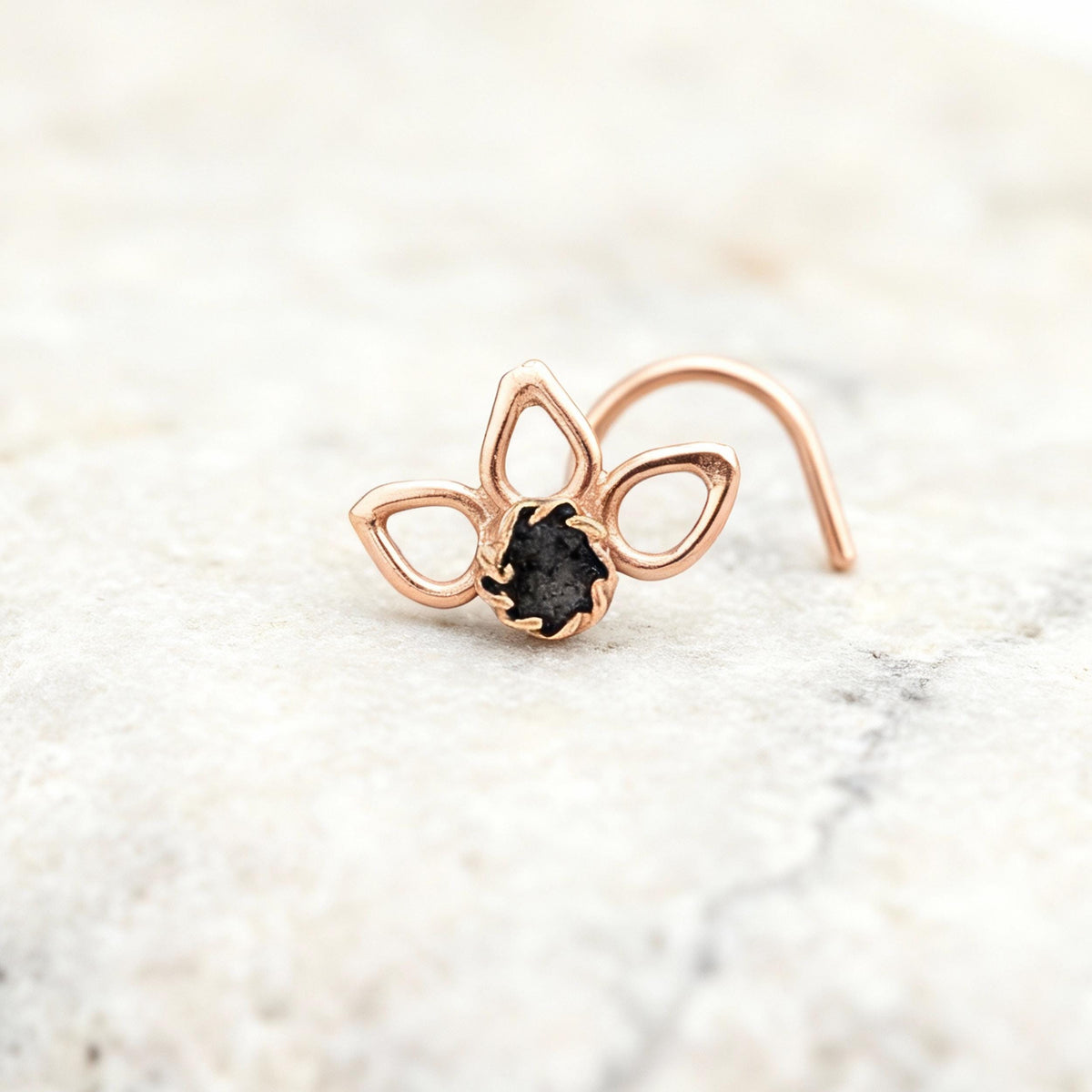 A rose gold nose ring with a black stone in the center is displayed on a white marble surface.