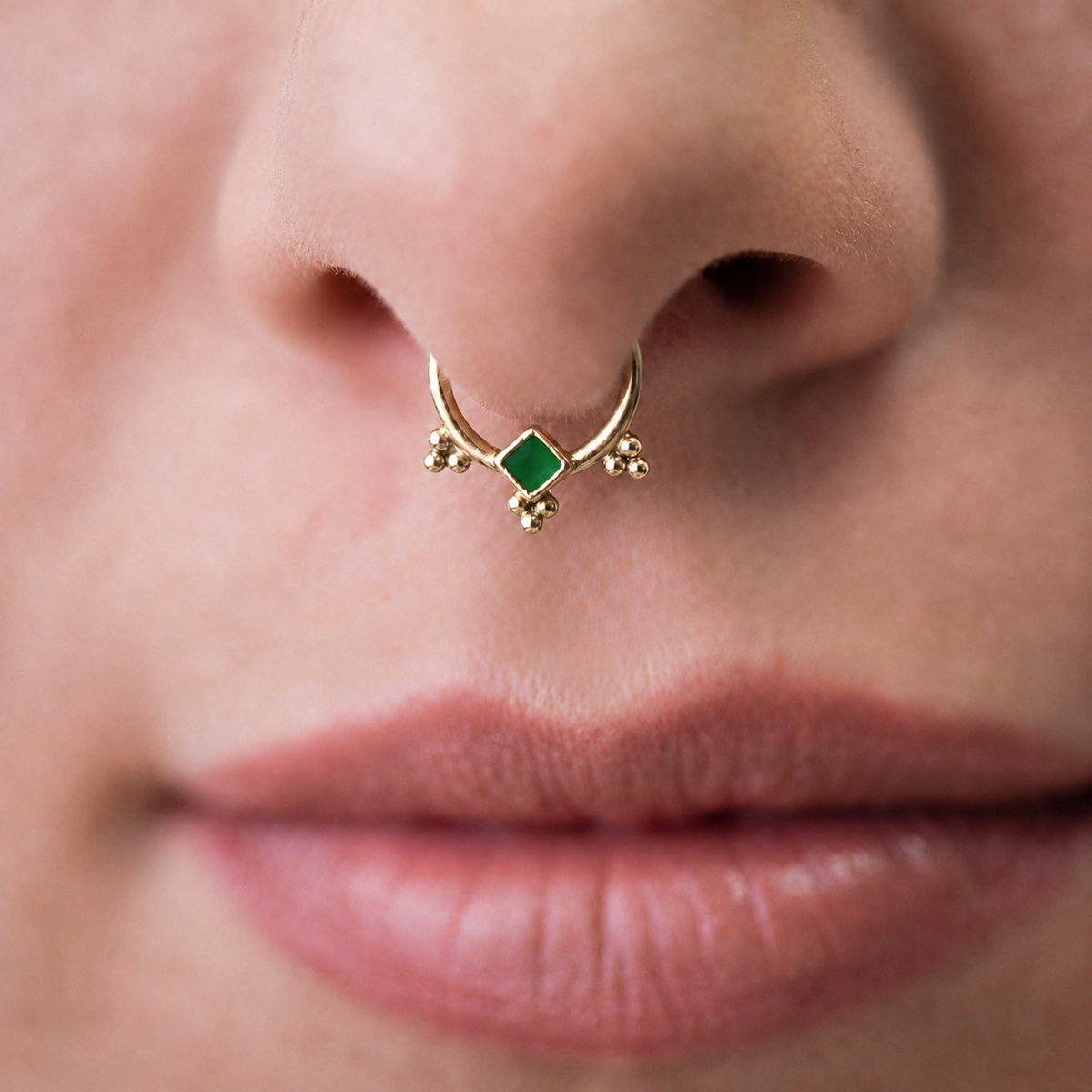 A close-up of a person's nose and lips, with a gold nose ring featuring a green gemstone in the center.