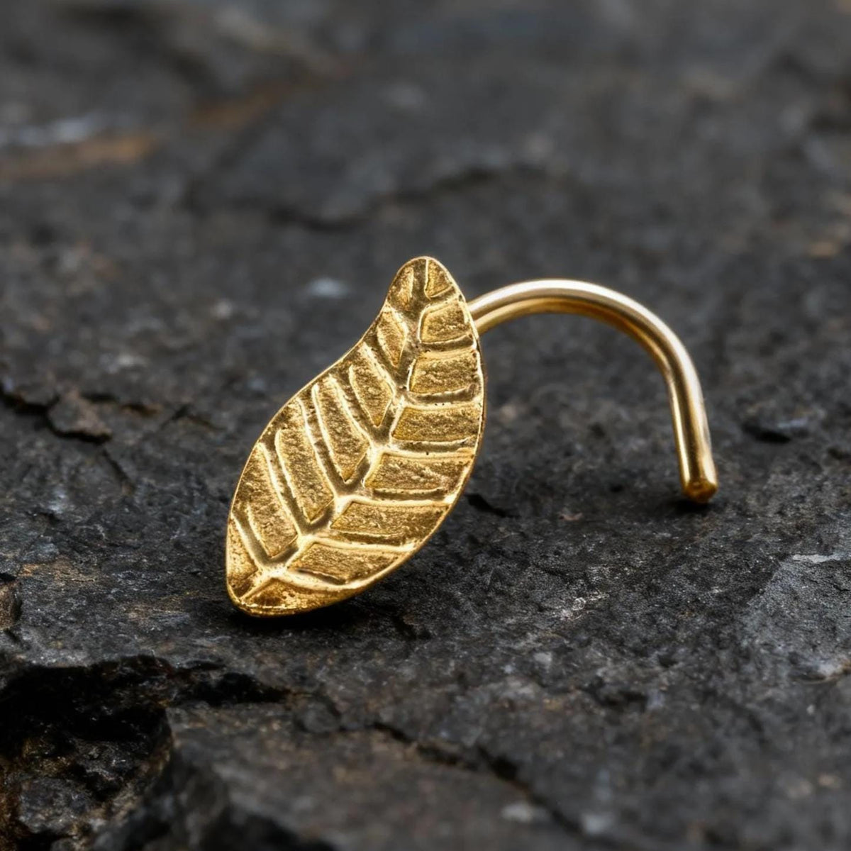 A gold leaf-shaped nose ring is resting on a dark, rocky surface.