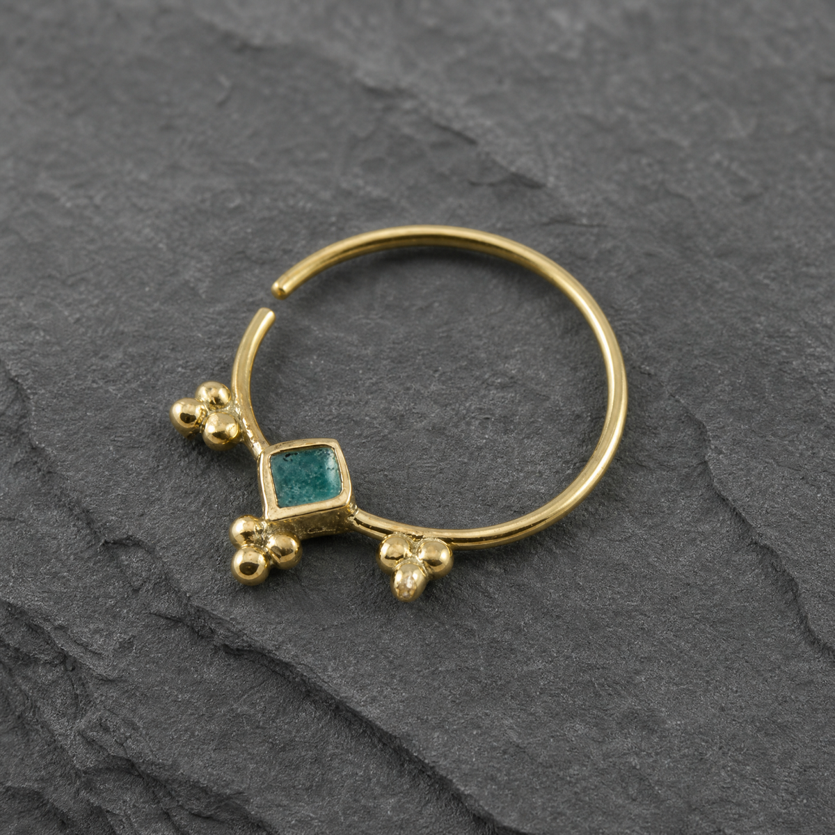 A gold-colored nose ring with a green gemstone is shown against a dark gray background.