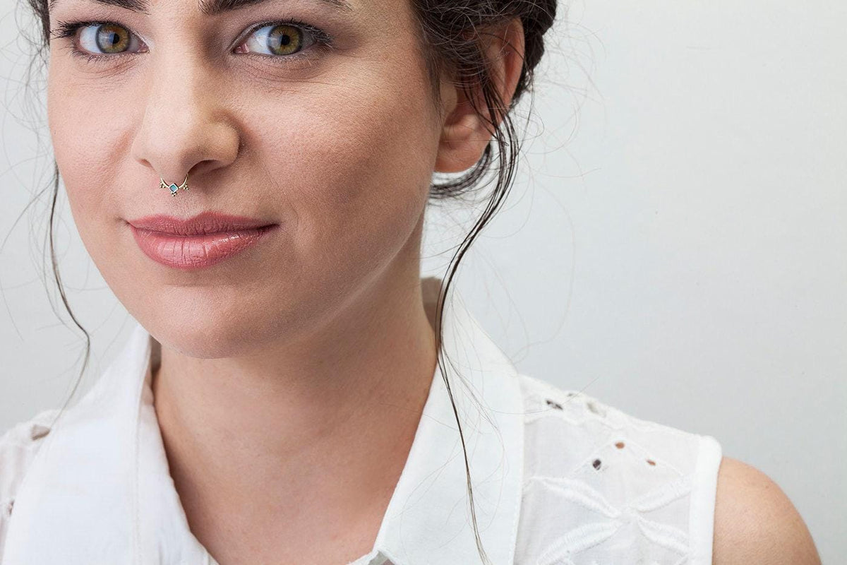A close-up portrait of a woman with a nose piercing, wearing a white sleeveless blouse with a floral pattern.