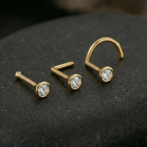 Three gold-colored nose studs with clear gemstones are displayed on a dark surface.