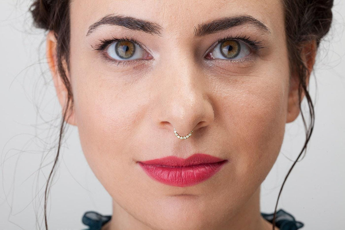 A close-up portrait of a woman with dark hair and striking red lipstick, wearing a nose ring.