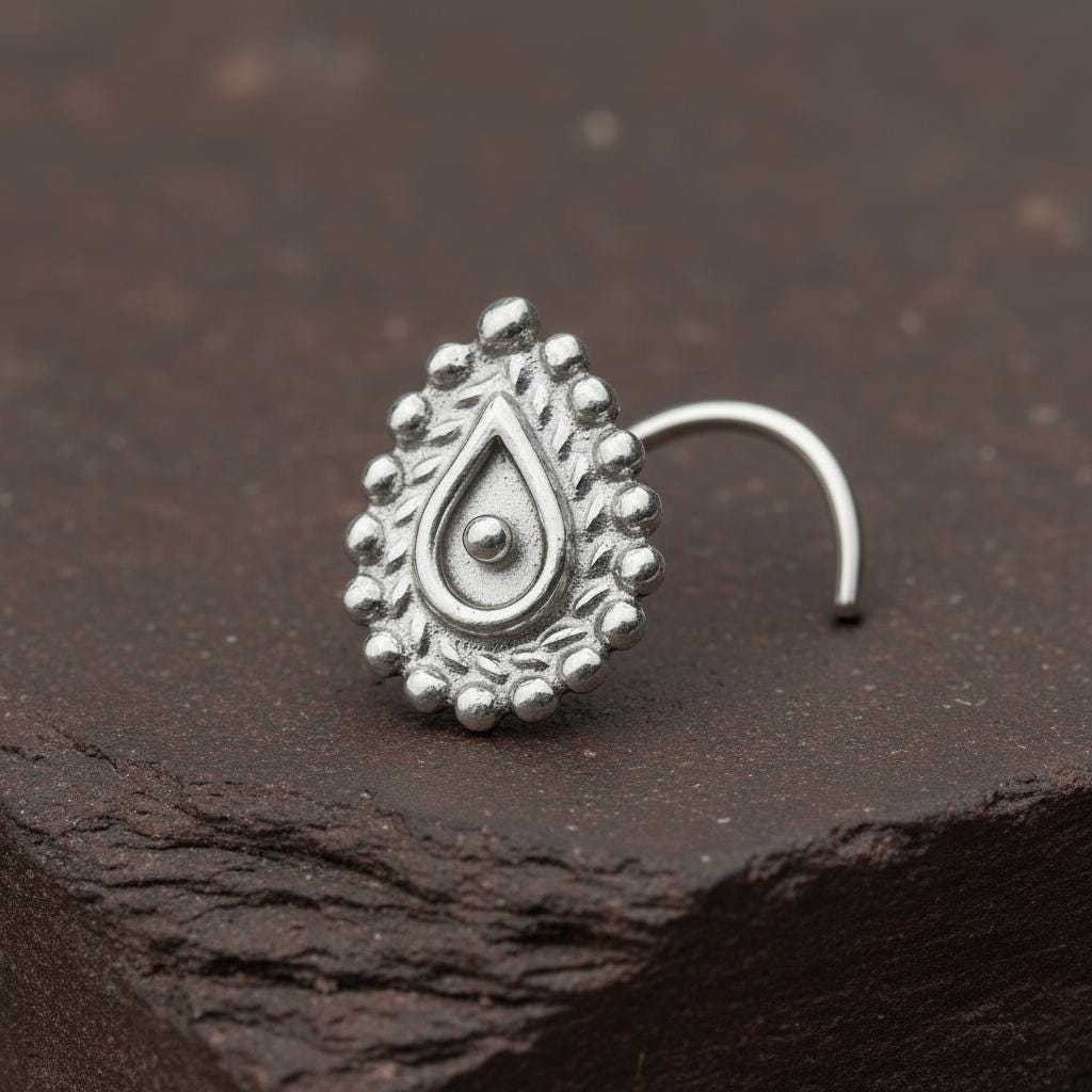 A silver nose ring with an ornate, teardrop-shaped design sits on a dark surface.