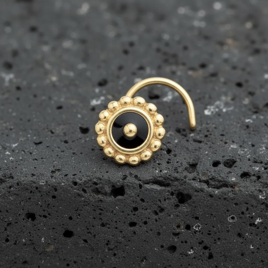 A gold-colored nose stud with a black center and a decorative pattern around it, resting on a dark surface.