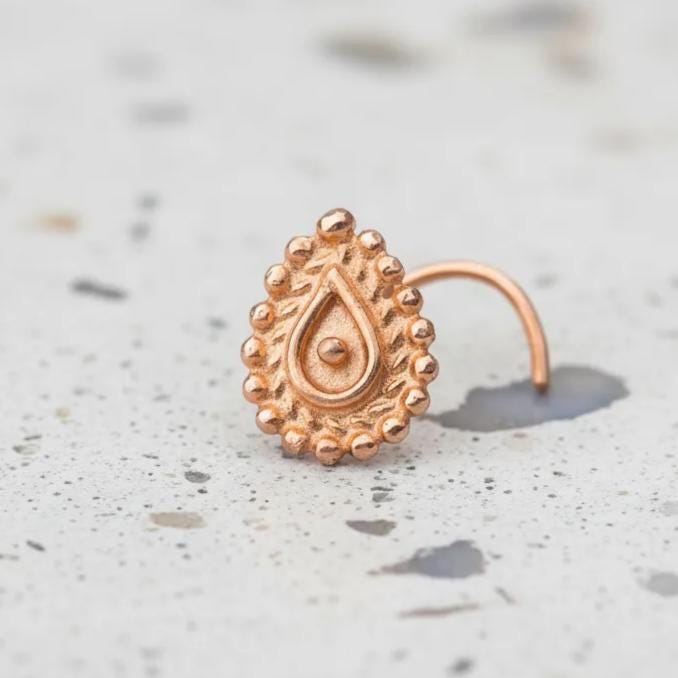 A close-up image of a gold-colored nose ring with an intricate design, resting on a speckled surface.