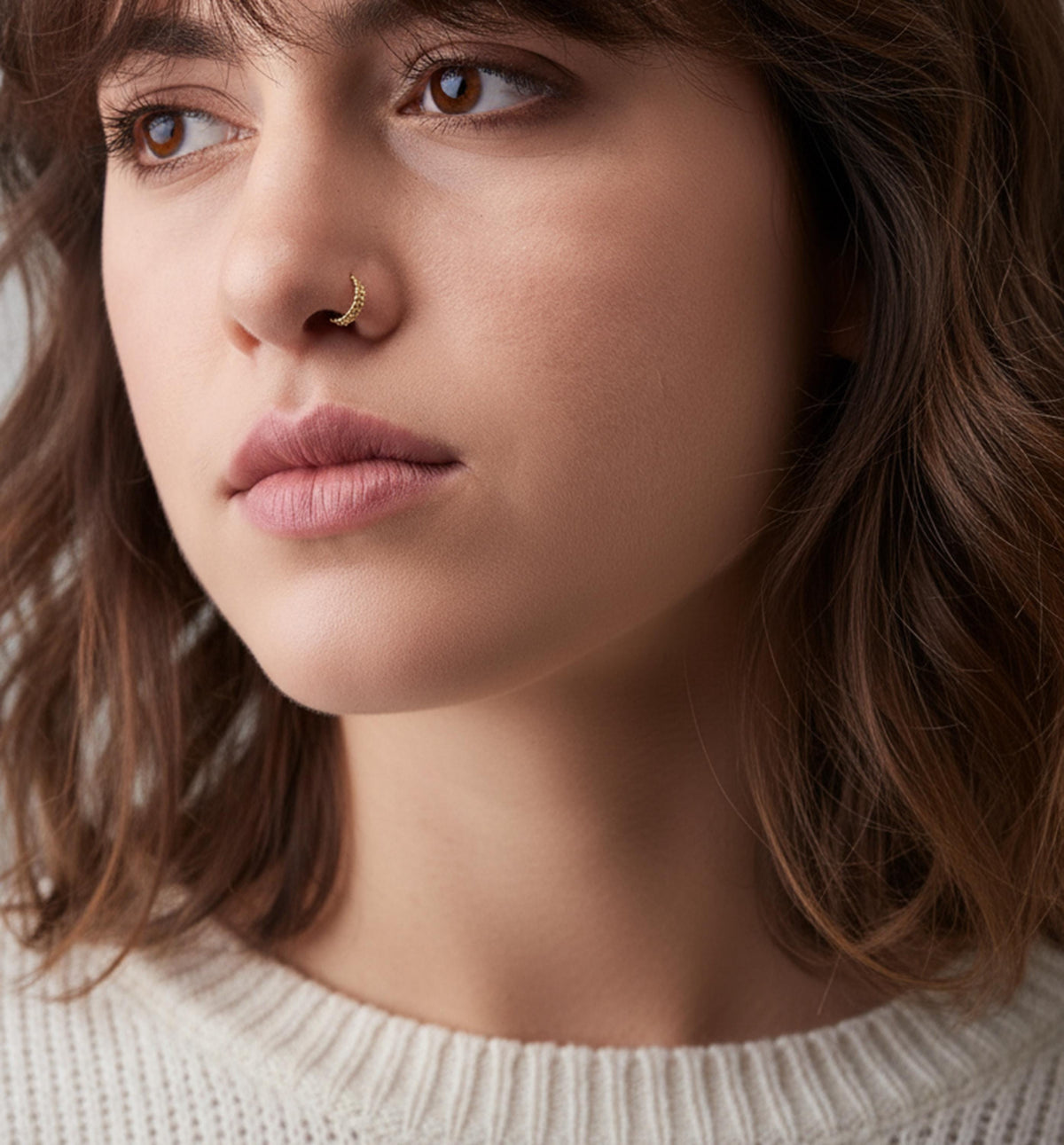 A close-up portrait of a young woman with brown hair, wearing a white sweater and a nose ring.