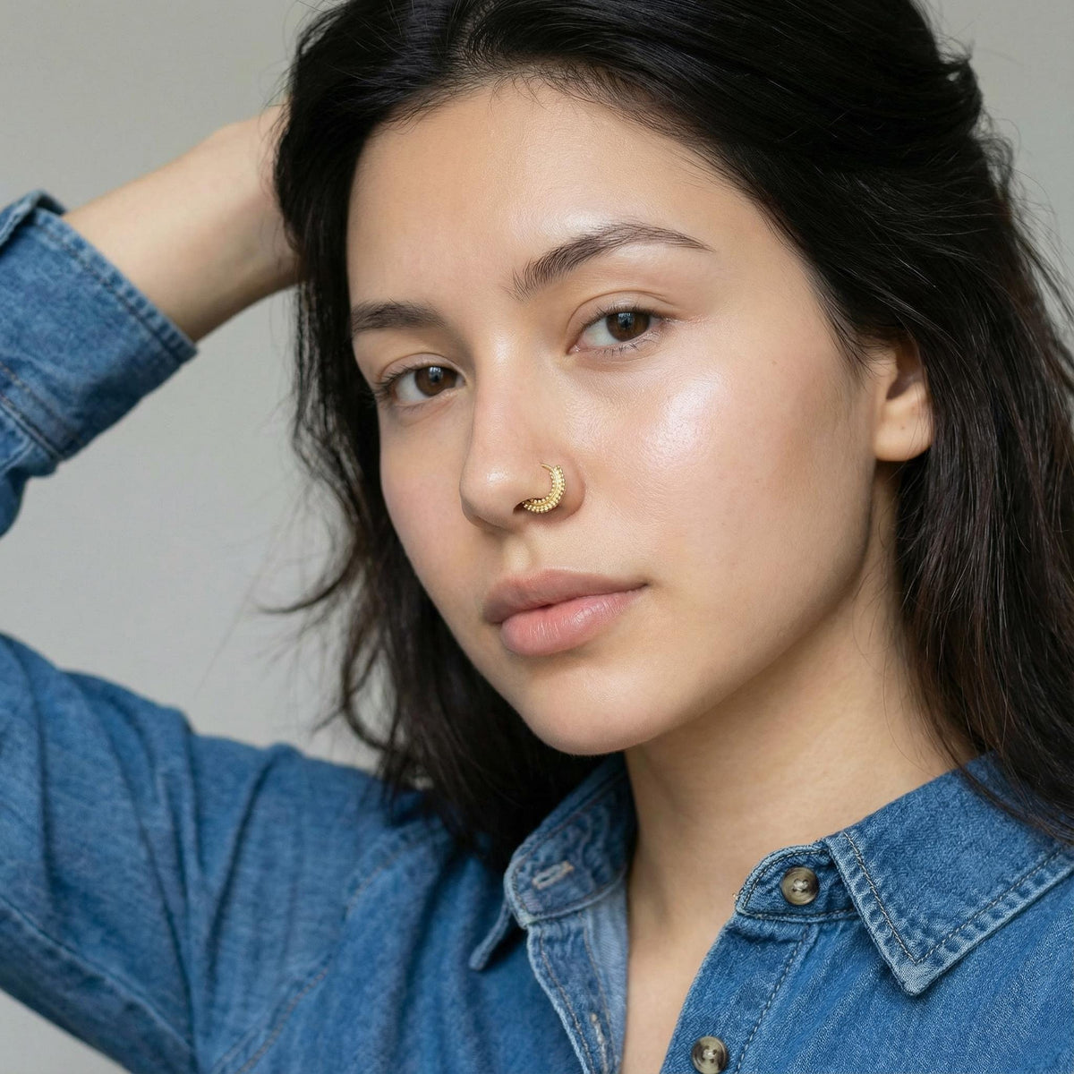 A close-up portrait of a young woman with dark hair, wearing a denim shirt and a nose ring.